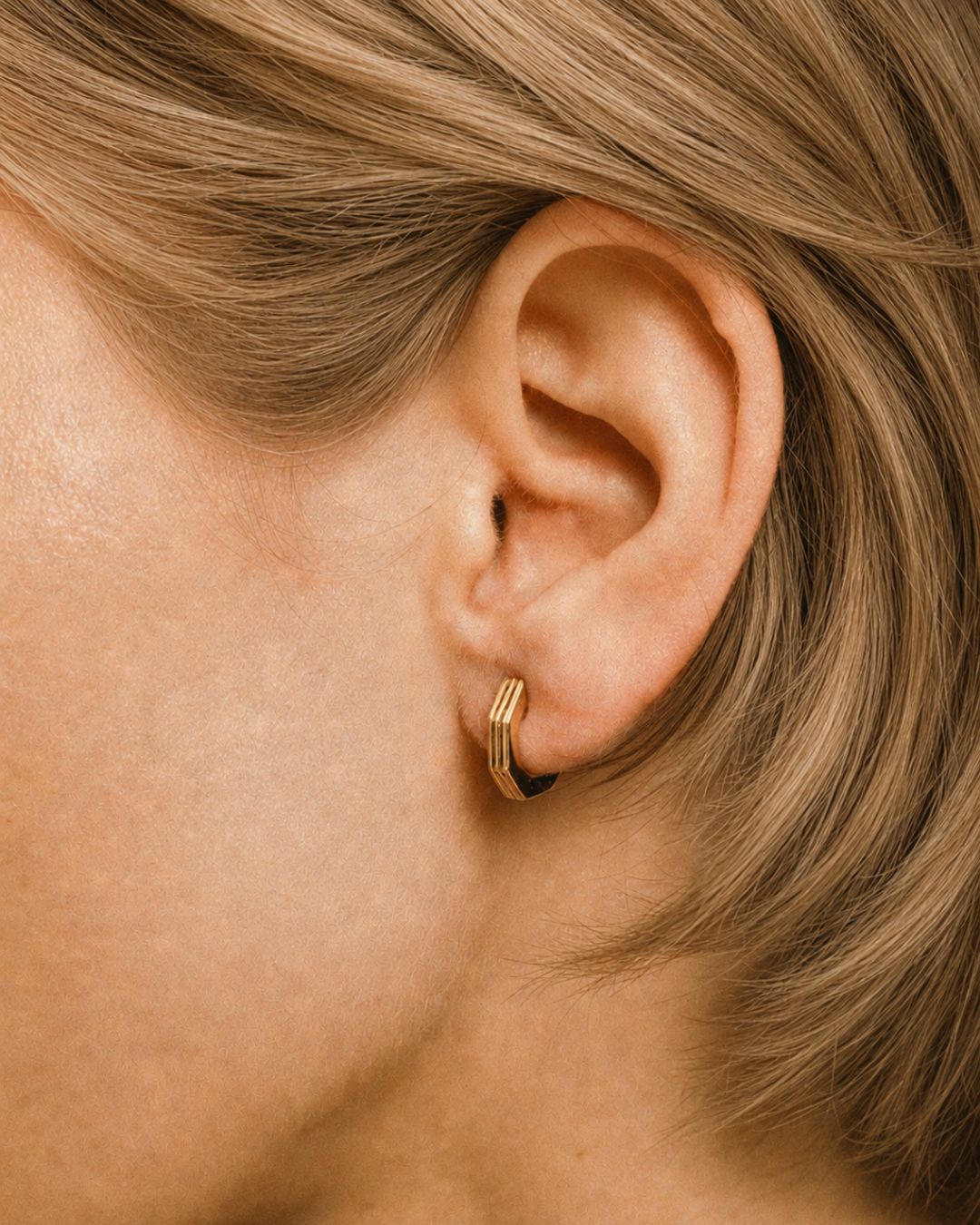 Close-up of an ear wearing a gold huggies earring with blonde hair.