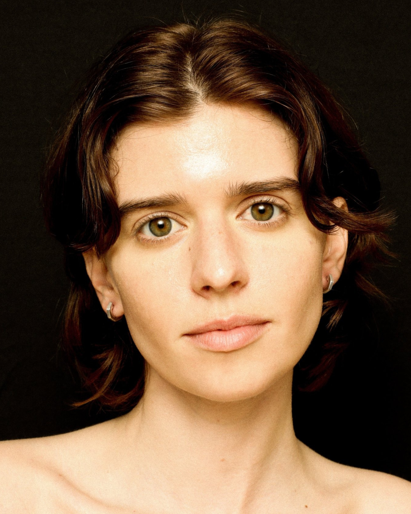 Close-up of a person with shoulder-length brown hair wearing silver geometric huggies earrings against a black background