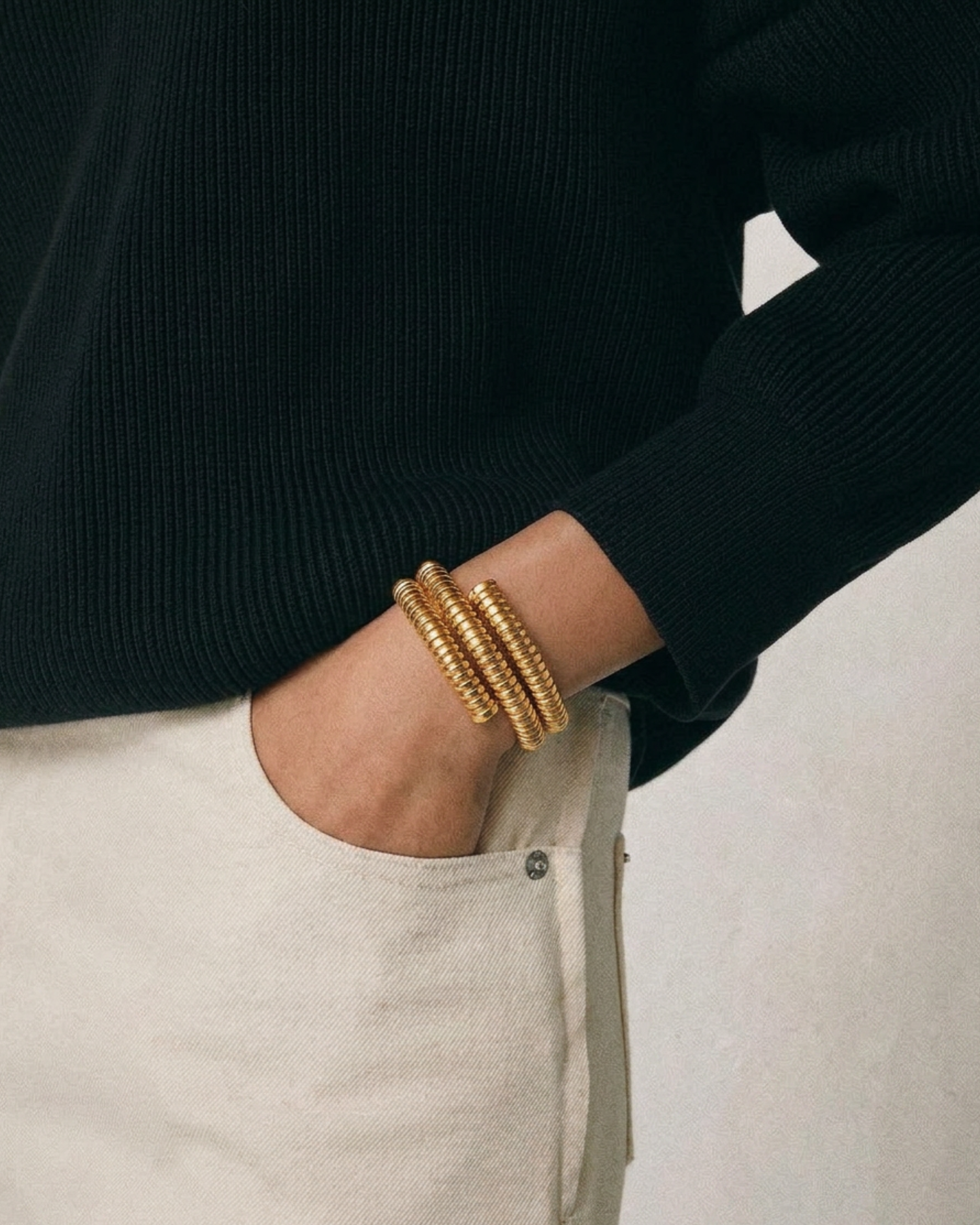 Person wearing a black sweater and beige pants with a gold bracelet on a neutral background