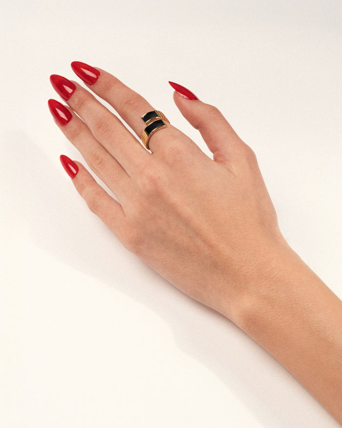 A gold-plated twist ring with polished black enamel stripes at the tips worn on a hand with red nails, shown in a lifestyle close-up.