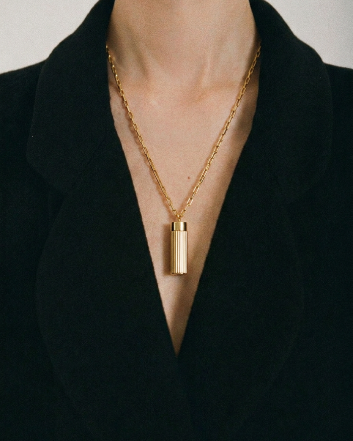 Gold necklace with a cylindrical pendant worn by a person against a black background
