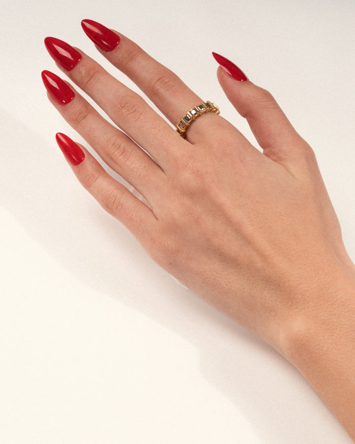 A gold-plated band ring with raised, squared frames set with shimmering pyrite stones worn on a hand with red nails, shown in a lifestyle close-up.