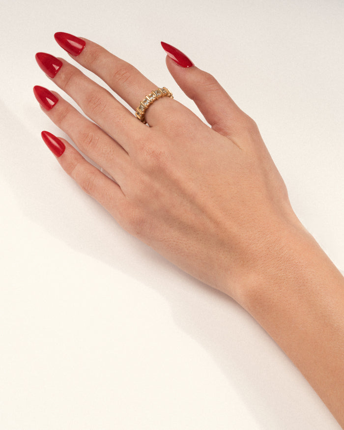 A gold-plated band ring with blue apatite stones set in raised, squared frames worn on a hand with red nails, shown in a lifestyle close-up.