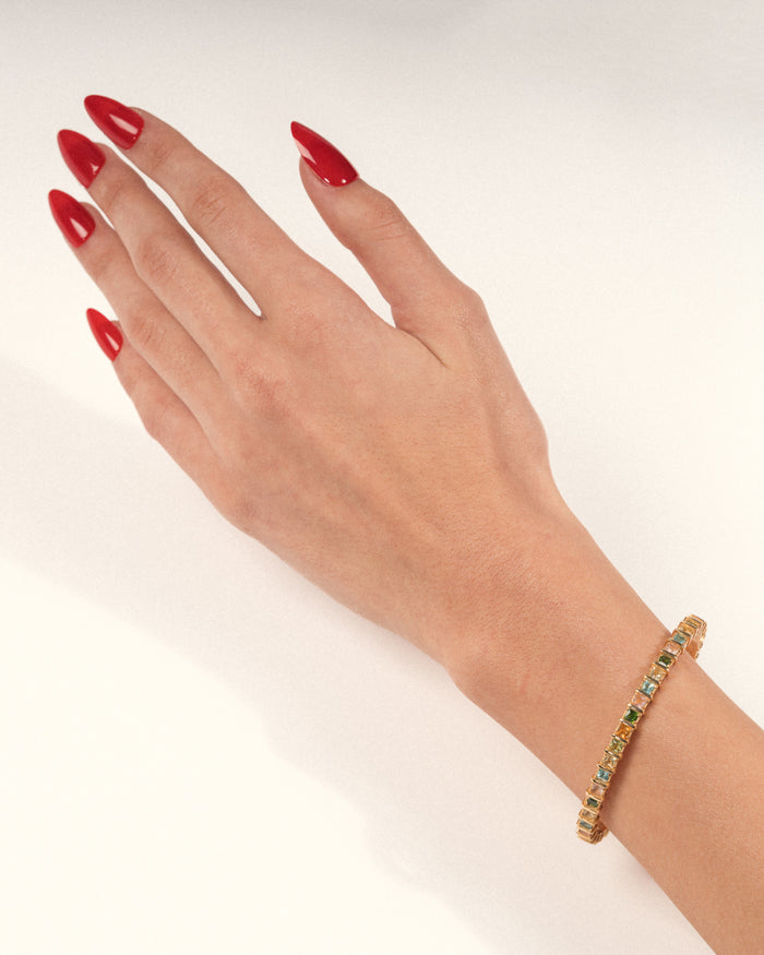 Gold-plated open cuff bracelet set with multicolored natural gemstones in raised square settings worn on a hand with red nails, shown in a lifestyle close-up.