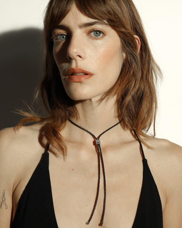A bolo tie with an adjustable silhouette crafted from braided leather, with signature rhodium-plated cubes at the fastening and tips worn by a model. 