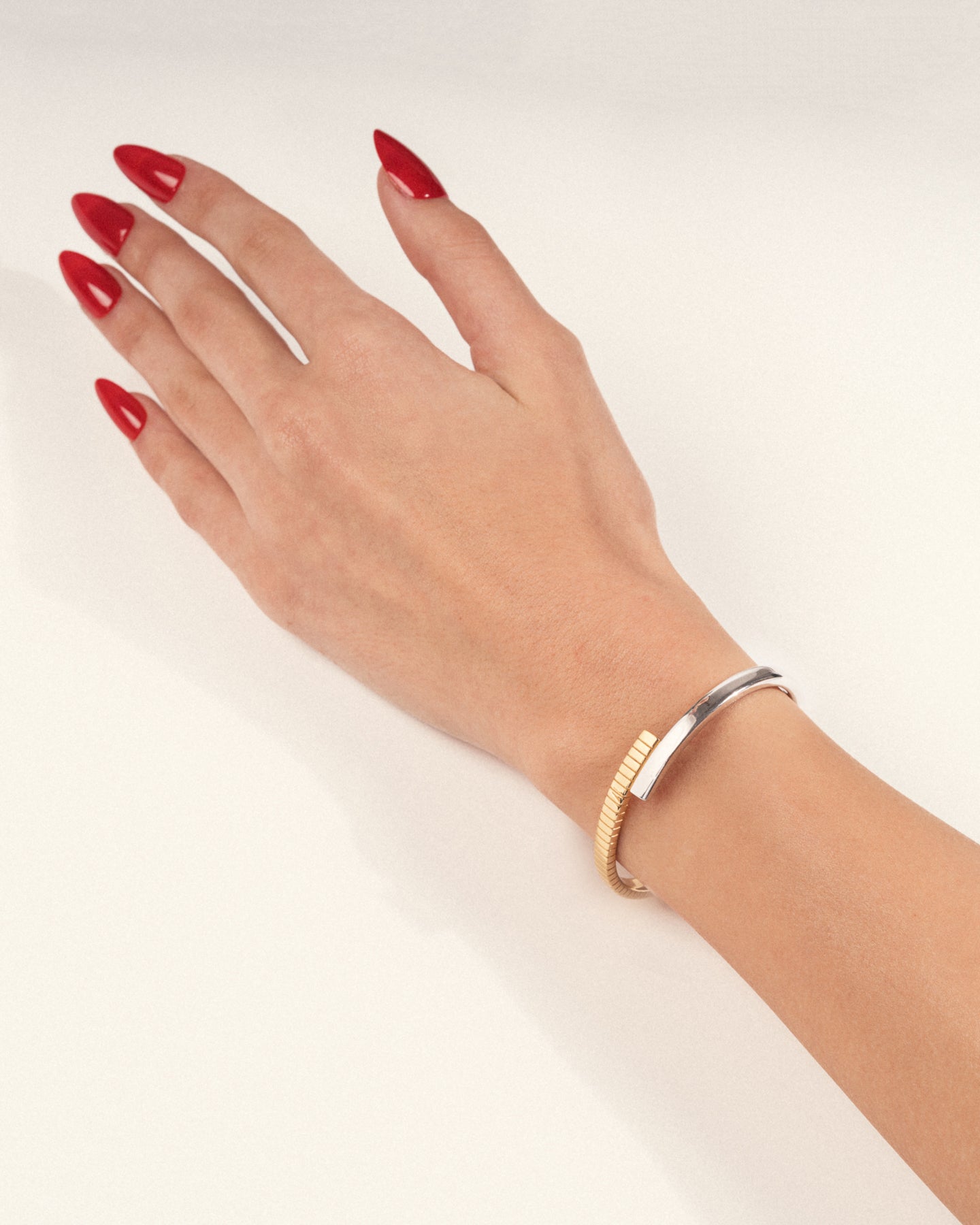 A two-tone (gold and silver) twist cuff bracelet with a mixed-metal finish worn on a hand with red nails, shown in a lifestyle close-up.