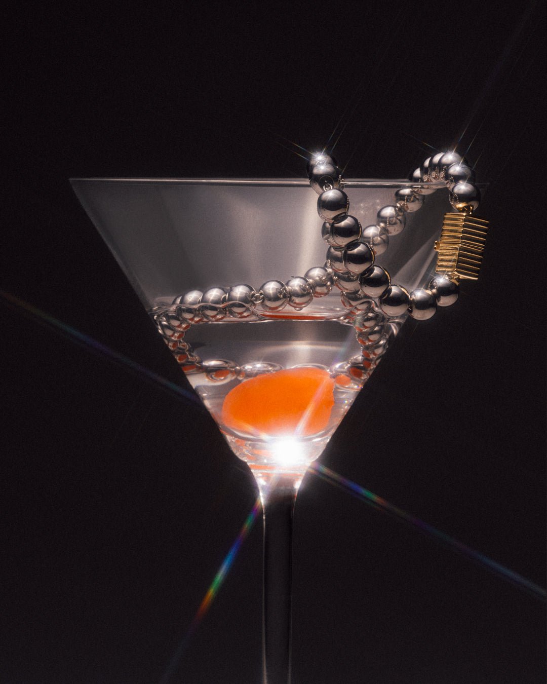 A silver necklace featuring a series of round rhodium-plated beads and a rectangular clasp with a small gold-tone detail placed in a martini glass. 