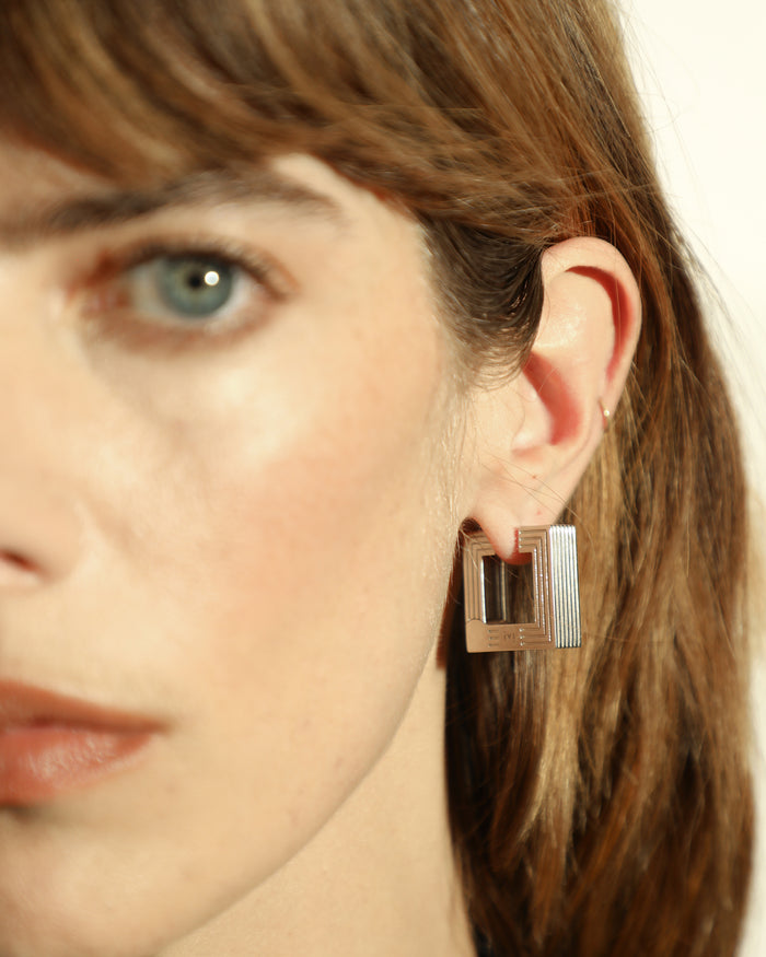 Pair of rhodium-plated medium Aurelia sculptural square hoop earrings worn by a model. 