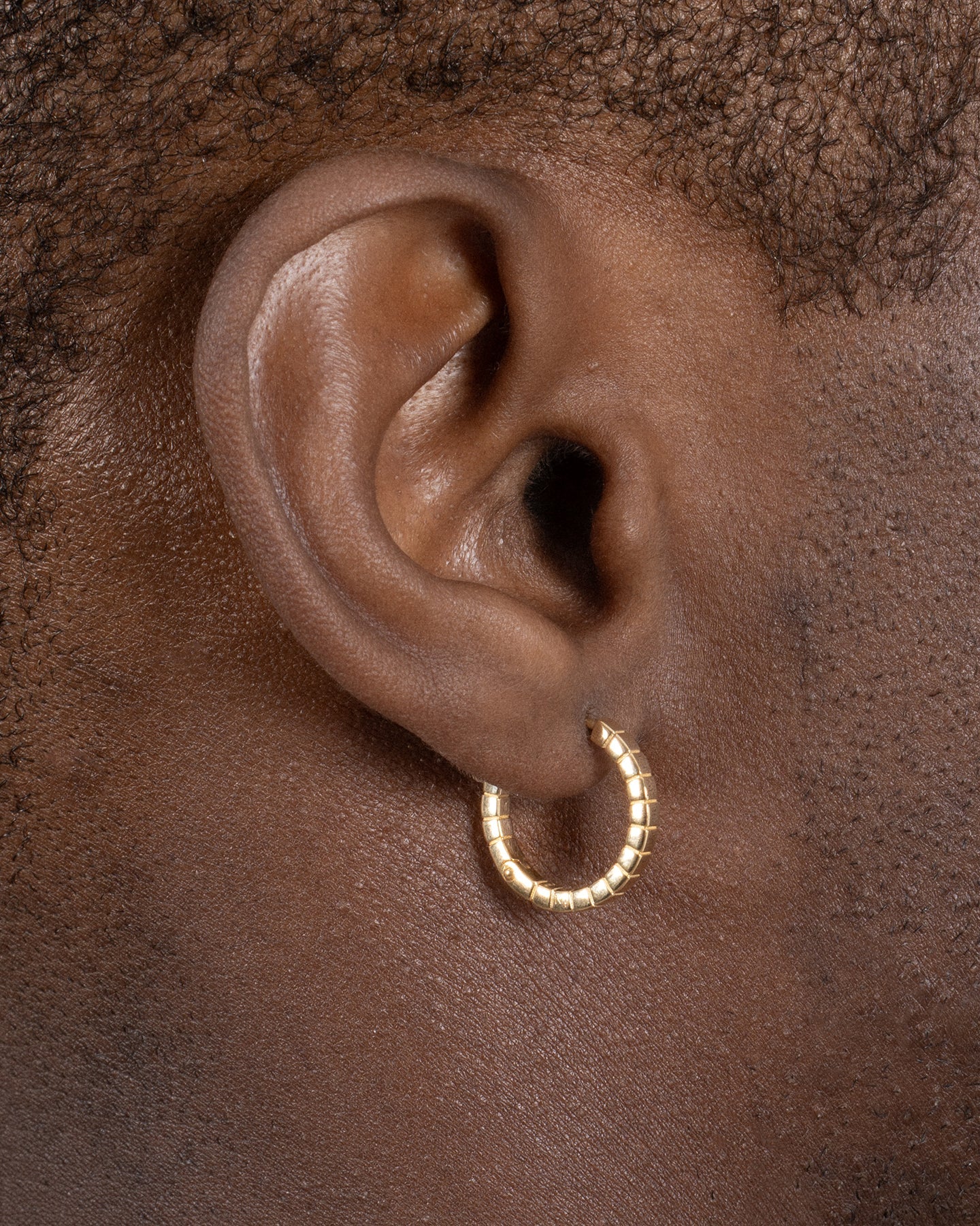 A pair of medium gold-plated Signore hoop earrings with a textured design, resembling a curving row of square beads worn by a model. 