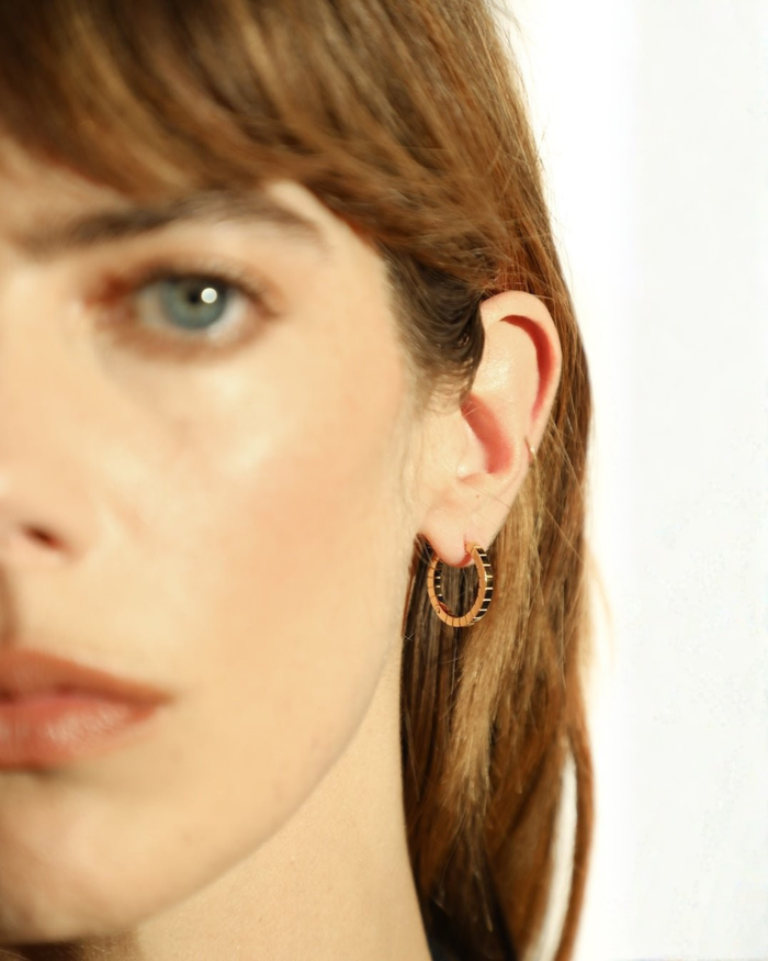 A pair of medium gold-plated Signore hoop earrings with a textured design, resembling a curving row of square beads worn by a model. 