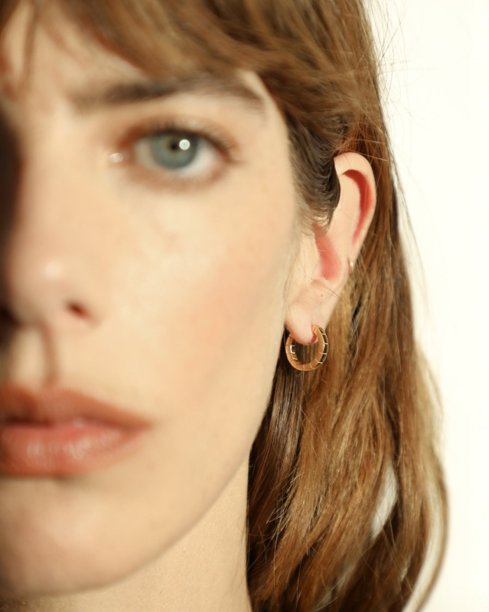 A pair of mini gold-plated Signore hoop earrings with a textured design, resembling a curving row of square beads worn by a model. 
