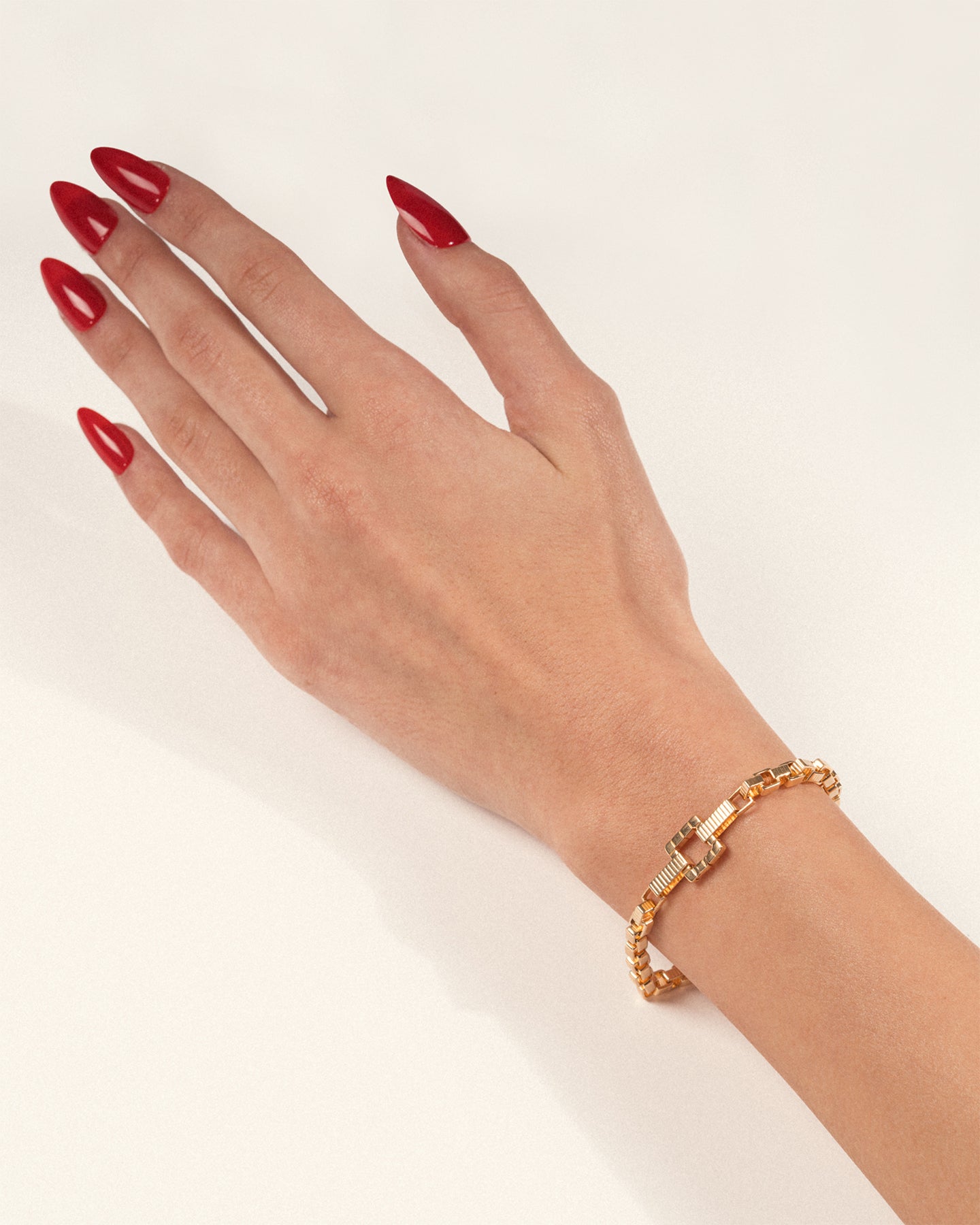 A gold-plated chain bracelet with angular links and a square clasp worn on a hand with red nails, shown in a lifestyle close-up.