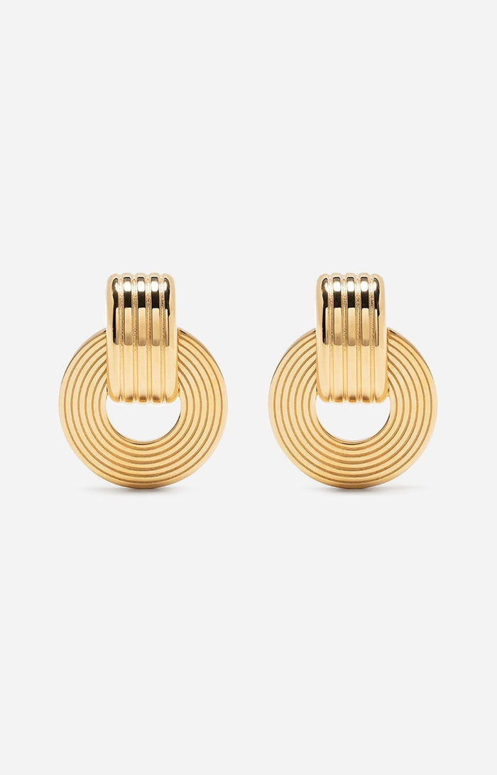 A pair of gold-plated oval hoop earrings with a circular pendant attached to the hoop, featuring intricate engravings.