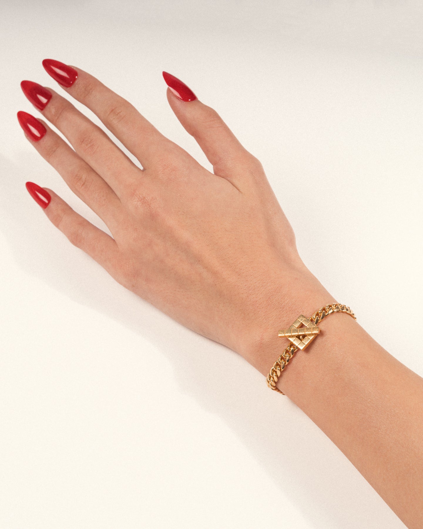 A gold-plated chunky chain necklace with a sculptural geometric clasp worn on a hand with red nails, shown in a lifestyle close-up.