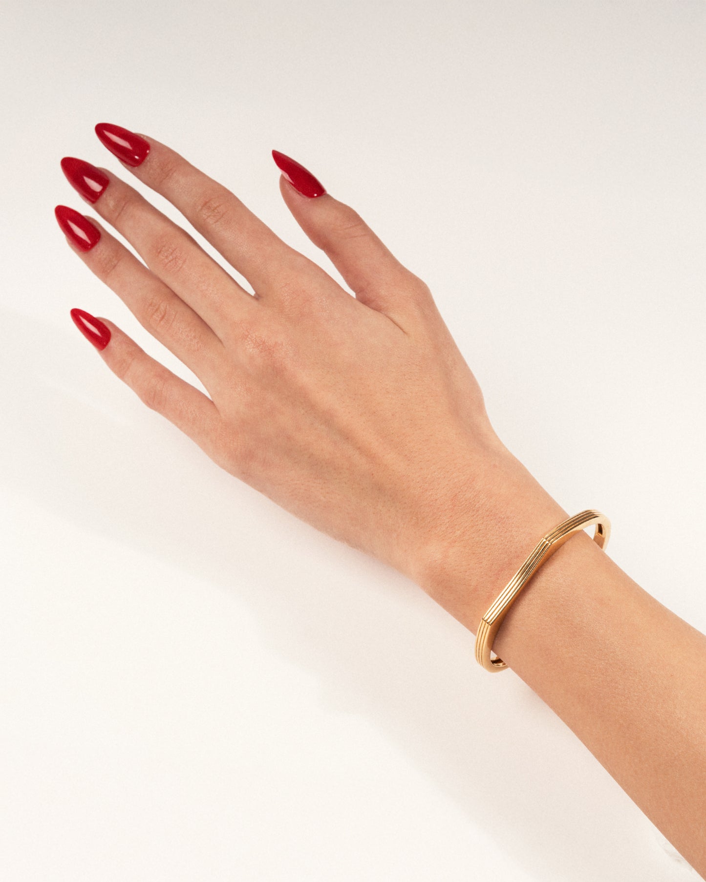 A gold-plated cuff bracelet with a sleek, modern design worn on a hand with red nails, shown in a lifestyle close-up.
