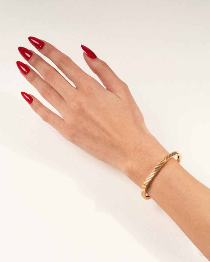 A gold-plated cuff bracelet with a sleek, modern design worn on a hand with red nails, shown in a lifestyle close-up.