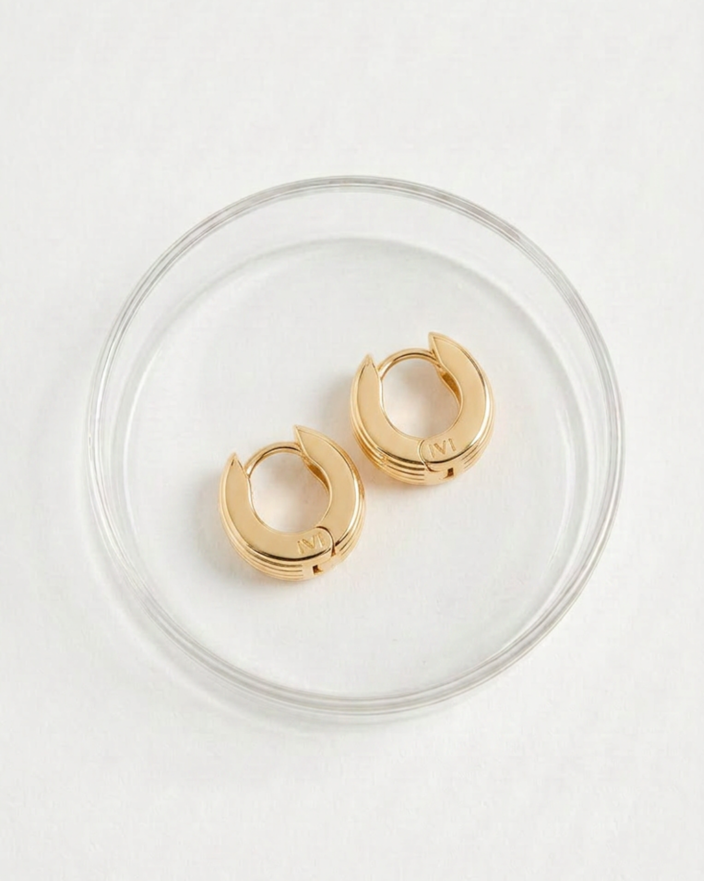 A pair of gold-plated round huggies earrings with a slim curved silhouette and engraved detailing placed in a glass packaging. 